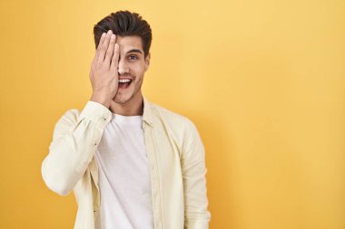 Young hispanic man standing over yellow background covering one eye with hand, confident smile on face and surprise emotion. 