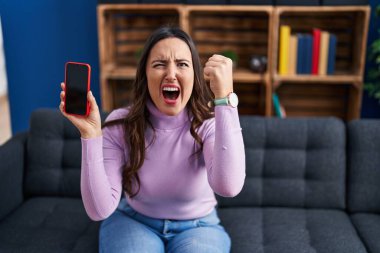 Young brunette woman holding smartphone showing blank screen annoyed and frustrated shouting with anger, yelling crazy with anger and hand raised 