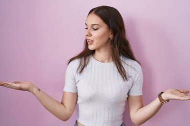 Pembe arka planda duran genç İspanyol kız gülümseyerek avuçlarını açık gösteriyor, sunuyor ve reklam karşılaştırması yapıyor ve dengeyi sağlıyor. 
