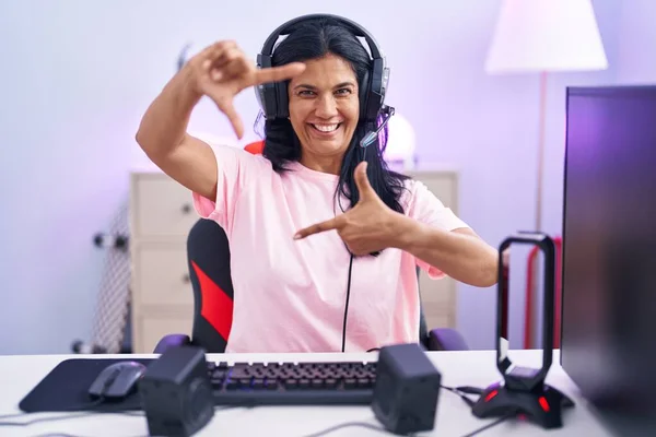 Mature hispanic woman playing video games at home smiling making frame with hands and fingers with happy face. creativity and photography concept.