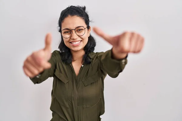 Hispanic woman with dark hair standing over isolated background approving doing positive gesture with hand, thumbs up smiling and happy for success. winner gesture.