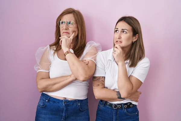 ヒスパニック系の母親と娘身に着けていますカジュアルな白いTシャツ上のピンクの背景思考心配質問 懸念と神経で手で顎 — ストック写真