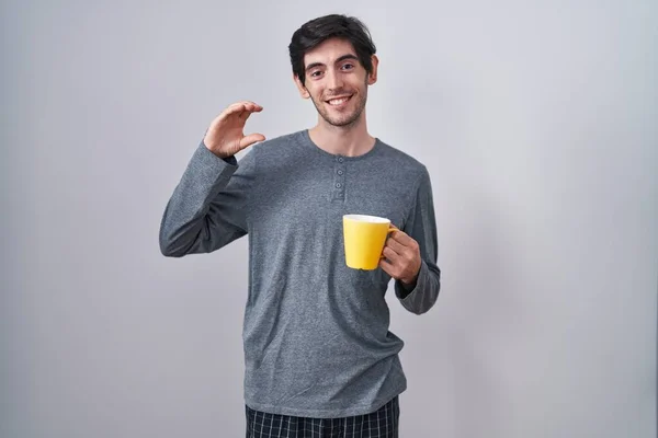 Young hispanic man wearing pajama drinking a cup of coffee smiling and confident gesturing with hand doing small size sign with fingers looking and the camera. measure concept.