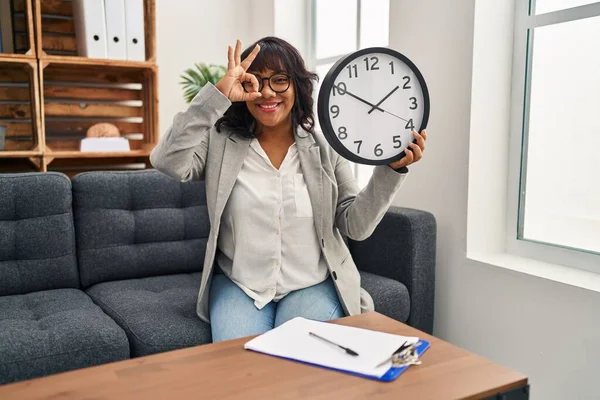 Hispanic woman working at therapy office holding clock smiling happy doing ok sign with hand on eye looking through fingers