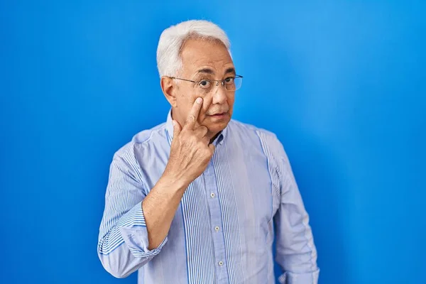 Hispanic senior man wearing glasses pointing to the eye watching you gesture, suspicious expression