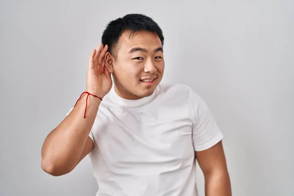 若い中国人男性の白い背景に手を耳に噂やゴシップへの聴聞会を聞いて笑みを浮かべて立っている 聴覚障害の概念 — ストック写真
