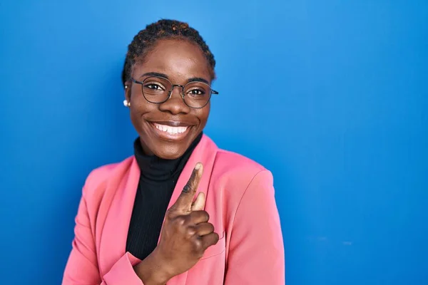 Beautiful black woman standing over blue background cheerful with a smile of face pointing with hand and finger up to the side with happy and natural expression on face
