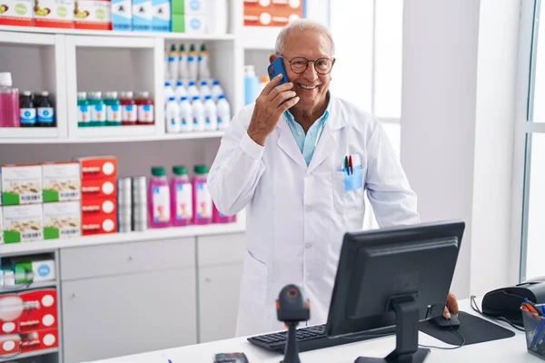 高级灰发男子药剂师在药店用电脑用智能手机交谈 — 图库照片
