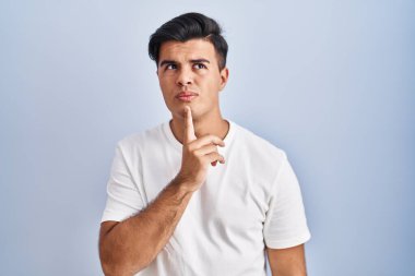 Hispanic man standing over blue background thinking concentrated about doubt with finger on chin and looking up wondering 