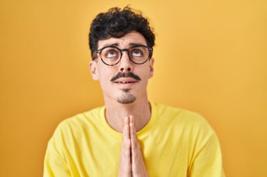 Sarı arka planda gözlük takan İspanyol bir adam elleri bağlı yalvarıyor ve dua ediyor. Yüzünde umut ifadesi var. Çok duygusal ve endişeli. Yalvarıyor. 