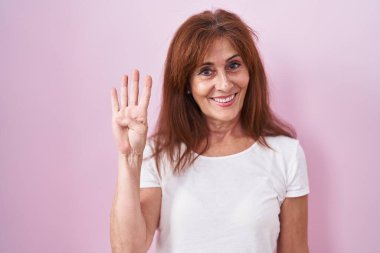 Pembe arka planda duran orta yaşlı bir kadın dört numarayı gösterip işaret ederken kendinden emin ve mutlu bir şekilde gülümsüyor.. 