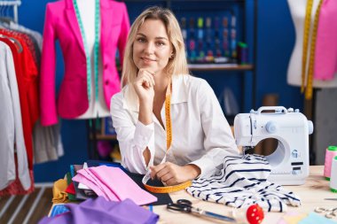 Young blonde woman tailor smiling confident sitting on table at tailor shop