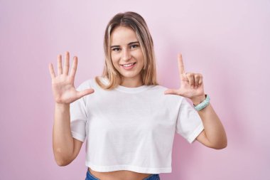 Pembe arka planda duran genç sarışın kadın yedinci parmaklarını gösterip işaret ederken kendinden emin ve mutlu bir şekilde gülümsüyor.. 