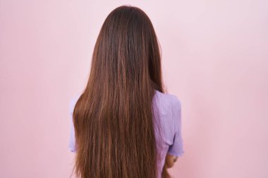 Pembe arka planda duran uzun saçlı genç İspanyol kadın çapraz kollarla arkasını dönüp bakıyor. 
