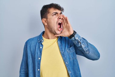 Young hispanic man standing over blue background shouting and screaming loud to side with hand on mouth. communication concept. 