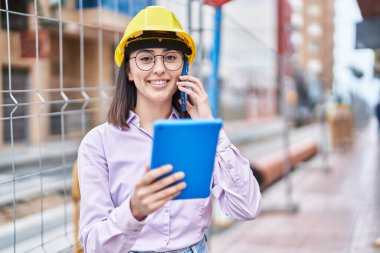 Young hispanic woman architect talking on the smartphone using touchpad at street
