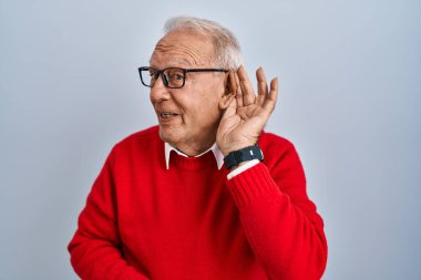 Senior man with grey hair standing over isolated background smiling with hand over ear listening an hearing to rumor or gossip. deafness concept. 