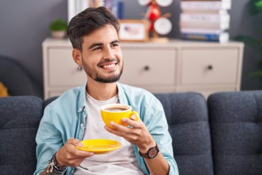 Genç İspanyol adam evdeki kanepede kahve içiyor.
