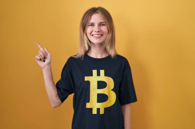 Sarışın, beyaz kadın bitcoin tişört giyiyor. Neşeli, yüzünde bir gülümseme var. Yüzünde mutlu ve doğal bir ifade var. 