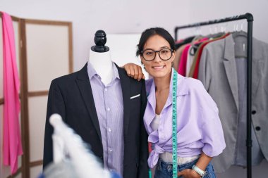Young beautiful hispanic woman tailor smiling confident leaning on manikin at tailor shop