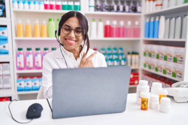 Eczanede çalışan genç, Arap bir kadın. Yüzünde mutlu ve doğal bir ifadeyle gülümseyen yüzü olan neşeli bir dizüstü bilgisayar kullanıyor. 