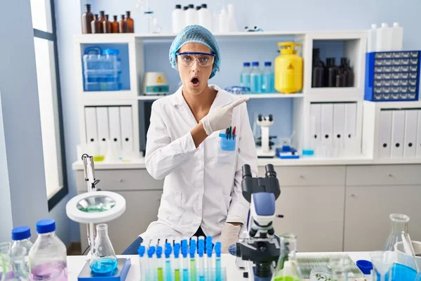 Brunette woman working at scientist laboratory surprised pointing with finger to the side, open mouth amazed expression.