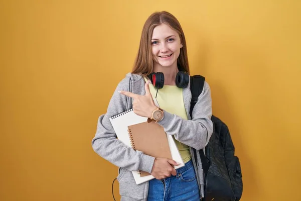 Young caucasian woman wearing student backpack and holding books cheerful with a smile on face pointing with hand and finger up to the side with happy and natural expression