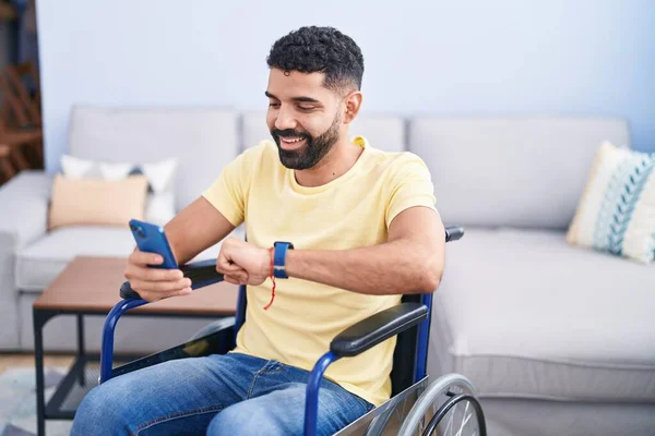 Young Arab Man Using Smartphone Looking Watch Sitting Wheelchair Home — Stok fotoğraf