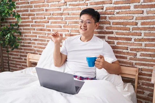 Young asian man drinking coffee sitting on the bed smiling happy pointing with hand and finger to the side
