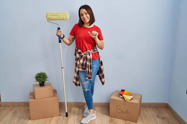 Young hispanic woman painting home walls with paint roller pointing to the back behind with hand and thumbs up, smiling confident