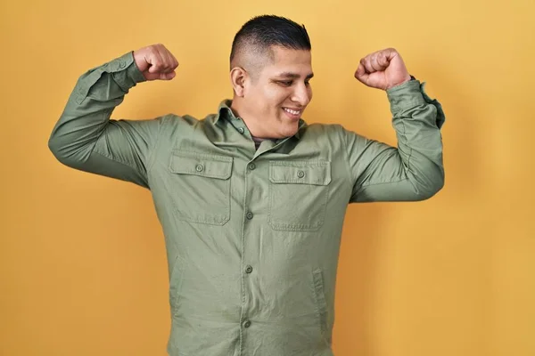 Jovem Hispânico Sobre Fundo Amarelo Mostrando Músculos Dos Braços Sorrindo — Fotografia de Stock