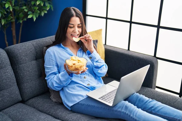 Young beautiful hispanic woman watching movie eating chips potatoes at home