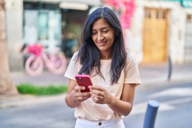 Genç, güzel, İspanyol bir kadın sokakta akıllı telefon kullanarak gülümsüyor.