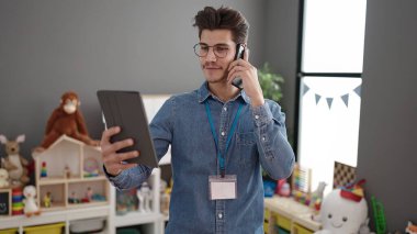 Young hispanic man preschool teacher using touchpad talking on smartphone at kindergarten