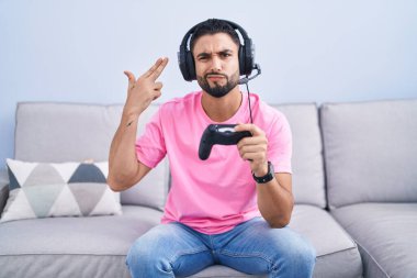 Hispanic young man playing video game holding controller sitting on the sofa shooting and killing oneself pointing hand and fingers to head like gun, suicide gesture.  clipart