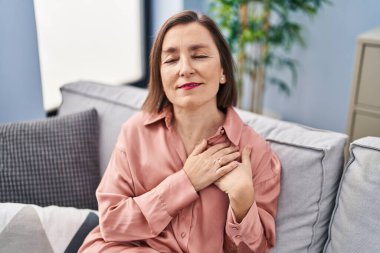 Middle age woman sitting on sofa with hands on heart at home