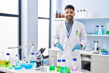 Young hispanic man wearing scientist uniform smiling confident at laboratory