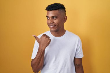Young hispanic man standing over yellow background smiling with happy face looking and pointing to the side with thumb up. 