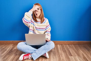 Evde dizüstü bilgisayar kullanan genç bir kadın yerde oturmuş şok içinde yüzünü ve gözlerini kapayarak utangaç ifadesiyle parmaklarının arasından bakıyor.. 