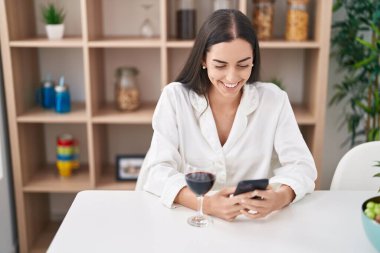 Young hispanic woman using smartphone and drinking wine at home