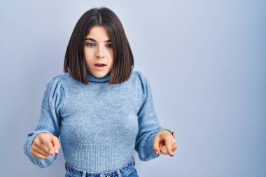 Young hispanic woman standing over blue background pointing down with fingers showing advertisement, surprised face and open mouth 