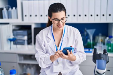 Laboratuvarda çalışan Latin Amerikalı genç bilim kadını akıllı telefon kullanıyor.