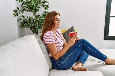 Evdeki kanepede kahve içen genç bir kadın.