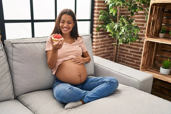 年轻的拉丁女人怀孕了 坐在沙发上吃着蛋糕 — 图库照片