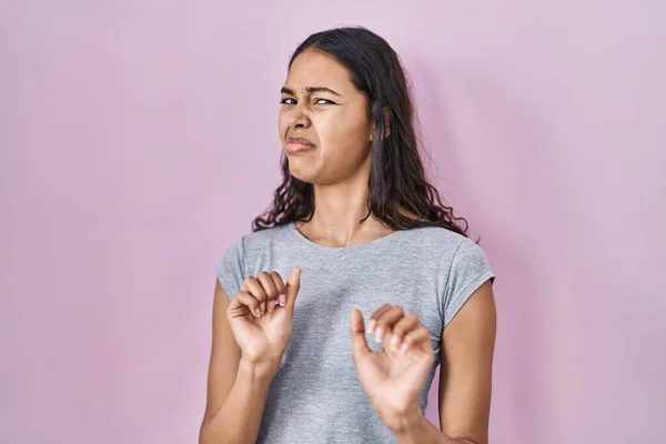 Young brazilian woman wearing casual t shirt over pink background disgusted expression, displeased and fearful doing disgust face because aversion reaction. with hands raised