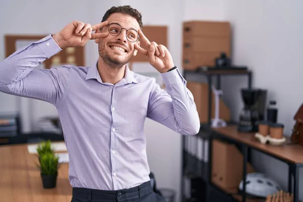 Young hispanic man at the office doing peace symbol with fingers over face, smiling cheerful showing victory