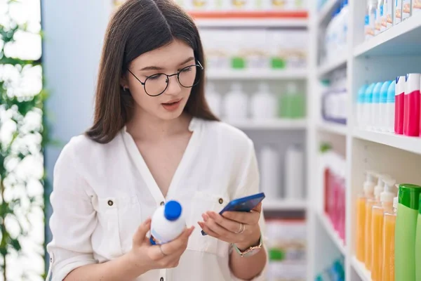 薬局で薬瓶を持っているスマートフォンを使用して若い白人女性の顧客 — ストック写真