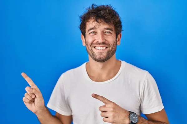西班牙裔年轻人站在蓝色的背景上微笑着 看着摄像机 用双手和手指指向旁边 — 图库照片