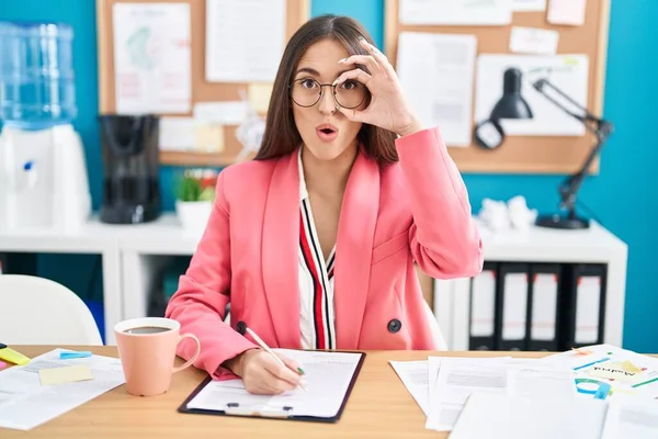 Young hispanic woman working at the office wearing glasses doing ok gesture shocked with surprised face, eye looking through fingers. unbelieving expression.