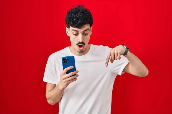 Spanyol Adam Kırmızı Arkaplan Üzerinde Akıllı Telefon Kullanıyor Parmaklarını Göstererek — Stok fotoğraf